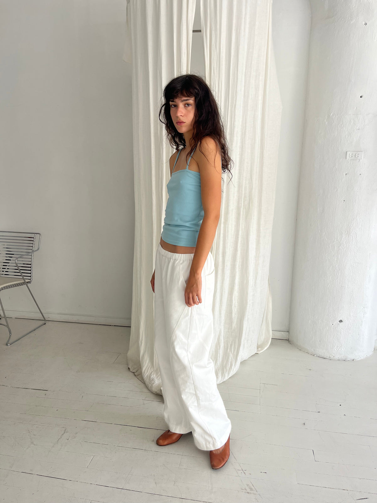 A woman stands in profile against a white curtain, wearing the Scallop Tank - Sky by Deiji Studios, a fitted ribbed tank with fine straps and scallop trim, styled with the Curve Pant in White and brown flats.