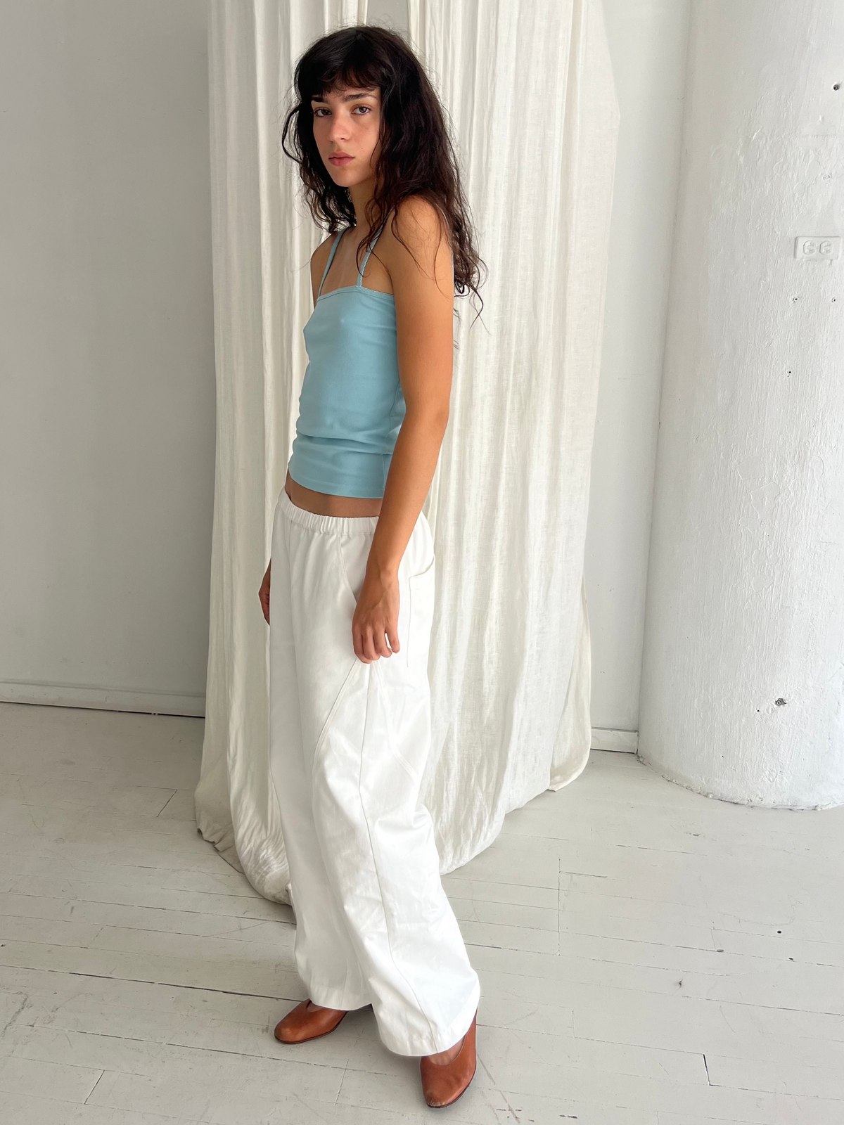 A woman with dark wavy hair stands in profile against a white curtain, wearing the Curve Pant - White by Deiji Studios, a softly structured pant with a relaxed fit and subtle curved leg, styled with the Scallop Tank in Sky.