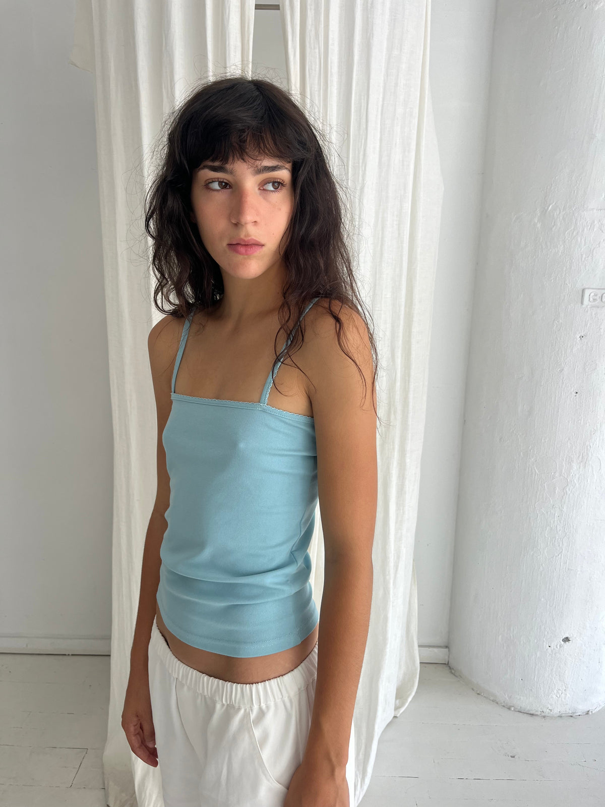 A woman with dark wavy hair stands against a white curtain, wearing the Scallop Tank - Sky by Deiji Studios, a fitted ribbed tank with fine straps and scallop trim, styled with the Curve Pant in White.