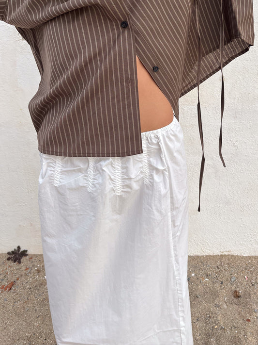 A person stands on sandy ground in front of a white wall, wearing the Channel Skirt and a brown, striped, partially unbuttoned shirt.
