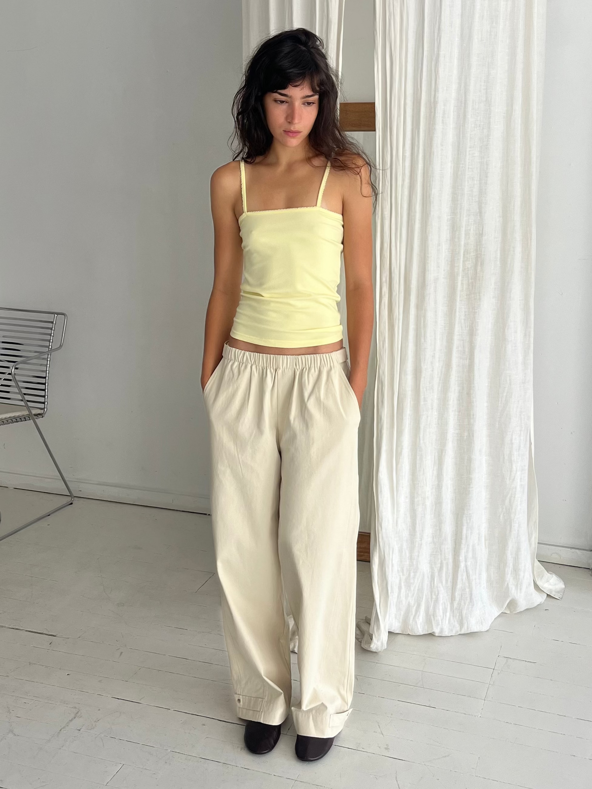 A woman with dark wavy hair stands against a white curtain, hands in her pockets, wearing the Scallop Tank - Lemon by Deiji Studios, a fitted ribbed tank with fine straps and scallop trim, with cream wide-leg pants.