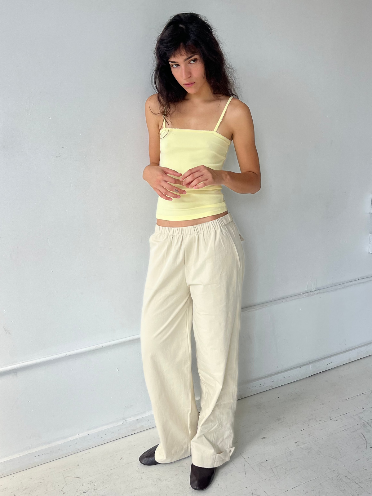 A woman with dark wavy hair stands against a white wall, wearing the Snap Tab Pant - Stone by Deiji Studios, a relaxed wide-leg pant with an elasticated waistband and snap tabs at the hem, styled with the Scallop Tank in Lemon.