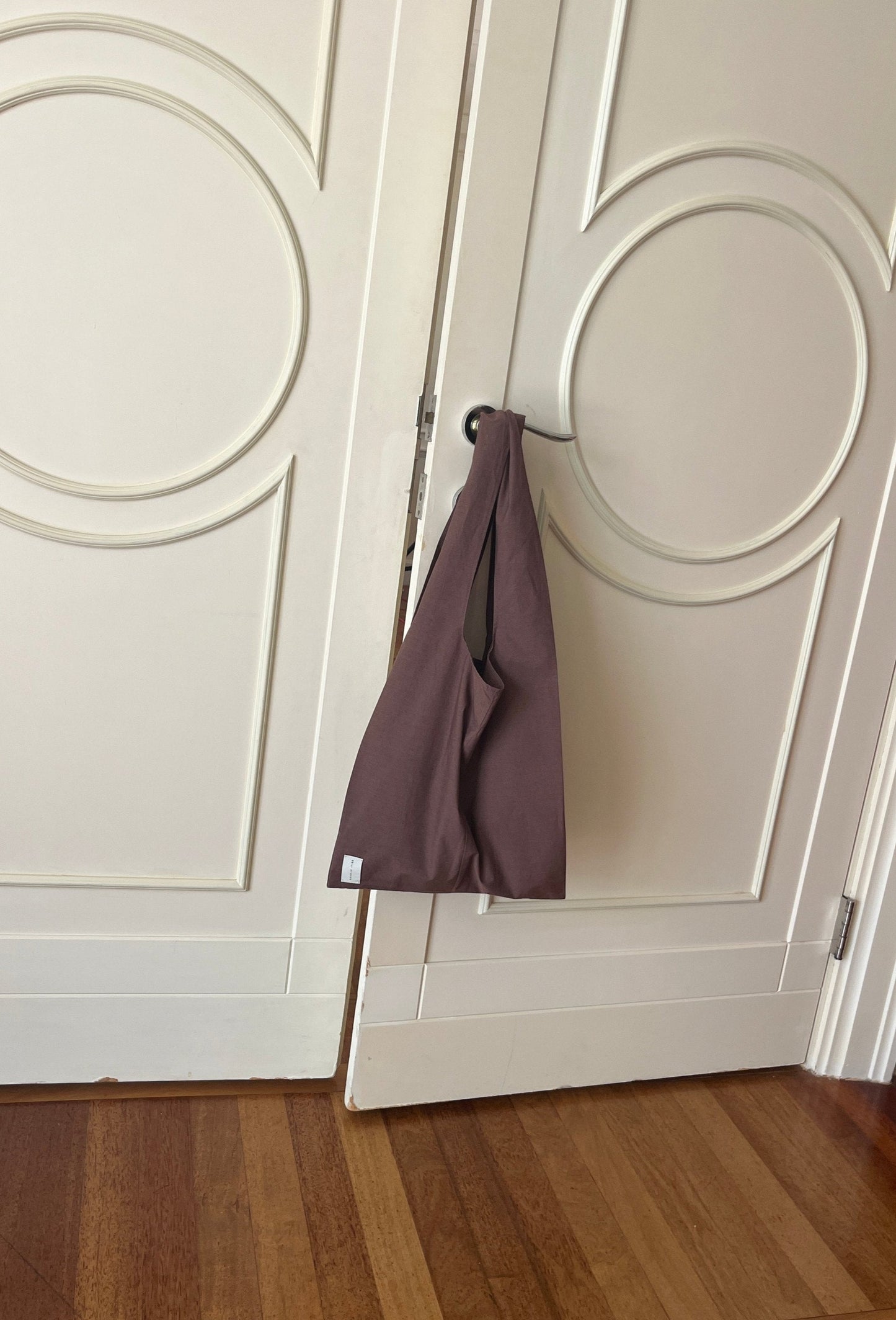 The Seam Tote, in mauve fabric, hangs on the handle of a white double door with circular designs, set against a wooden floor.
