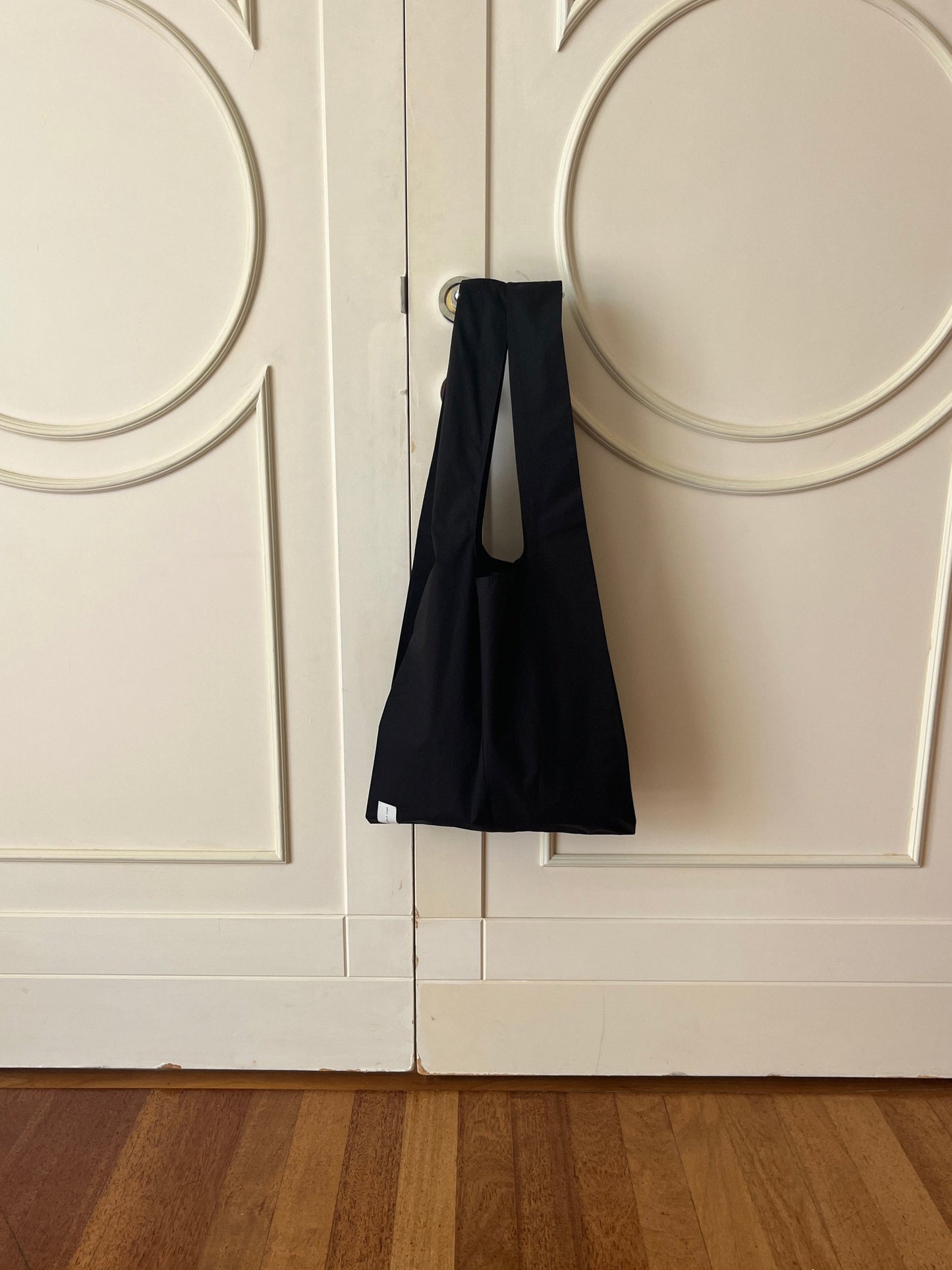 The Seam Tote - Black from Deiji Studios hangs from a round doorknob on a closed white double door with circular designs, above a wooden floor.