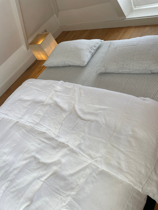 A neatly made bed features white linens from the Duvets collection and two striped pillows in a bright room with wooden floors. A small cube-shaped lamp sits by the wall, and sunlight streams in through a nearby window.