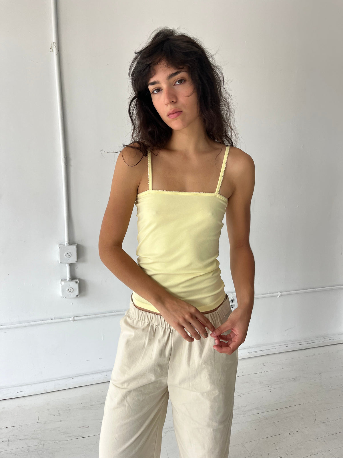 A woman stands against a white wall, wearing the Scallop Tank - Lemon by Deiji Studios, a fitted ribbed tank with fine shoulder straps and scallop trim along the straight neckline, with cream wide-leg pants.
