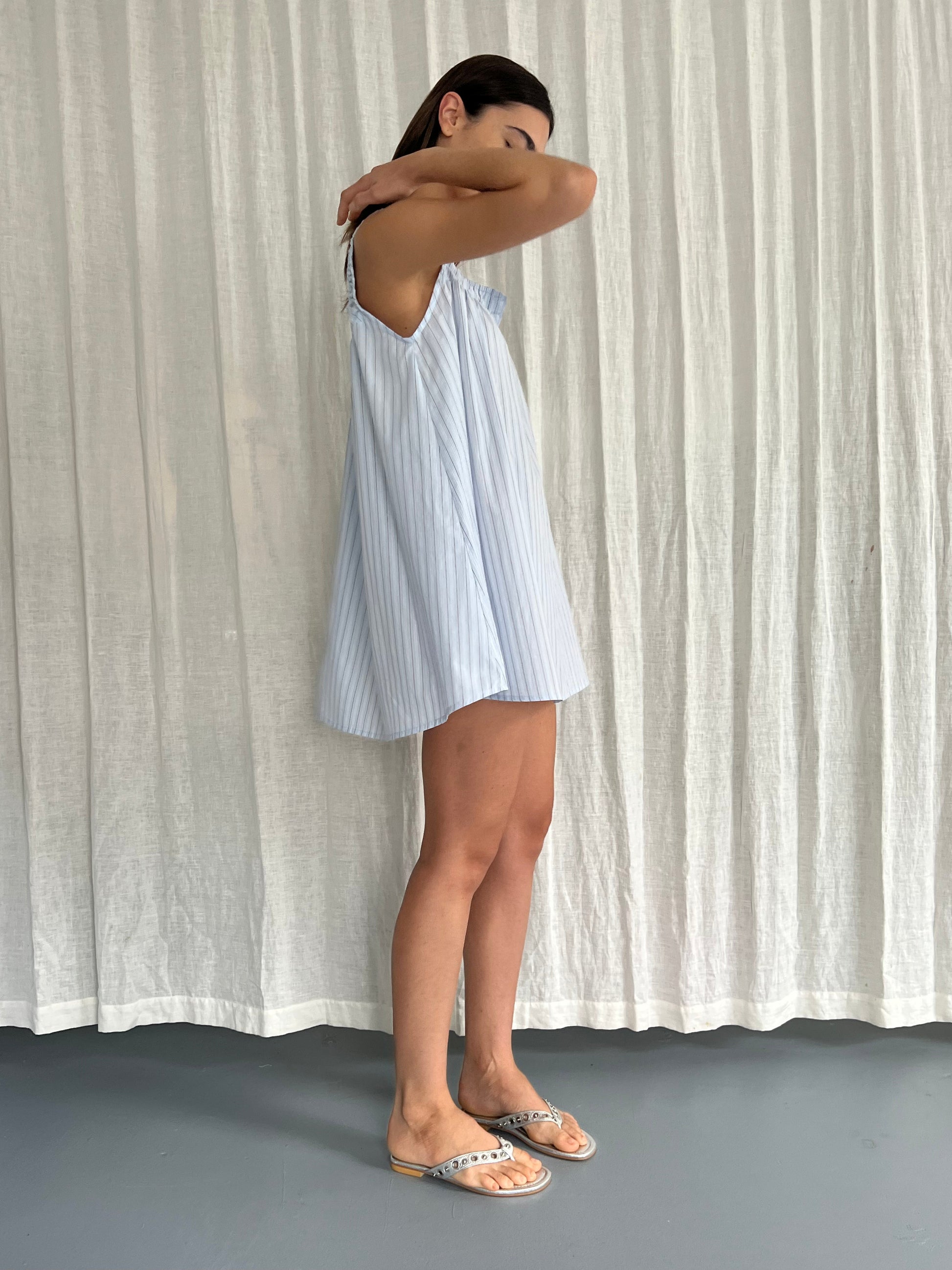 A woman in a light blue, sleeveless Yoke Mini Dress - Mineral Stripe from Deiji Studios, crafted from organic cotton, stands sideways before a white curtain with arms raised and face partially hidden, wearing flat, open-toe sandals.