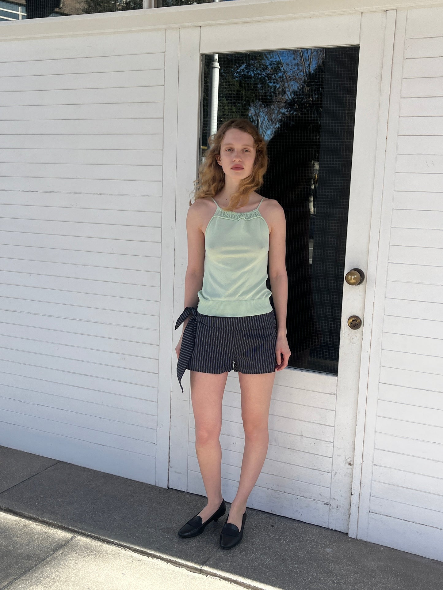 A woman with wavy blonde hair stands before a white door on a sunny day, wearing the mint Drawcord Knit Top by Deiji Studios, navy striped side-tie shorts, and black loafers.