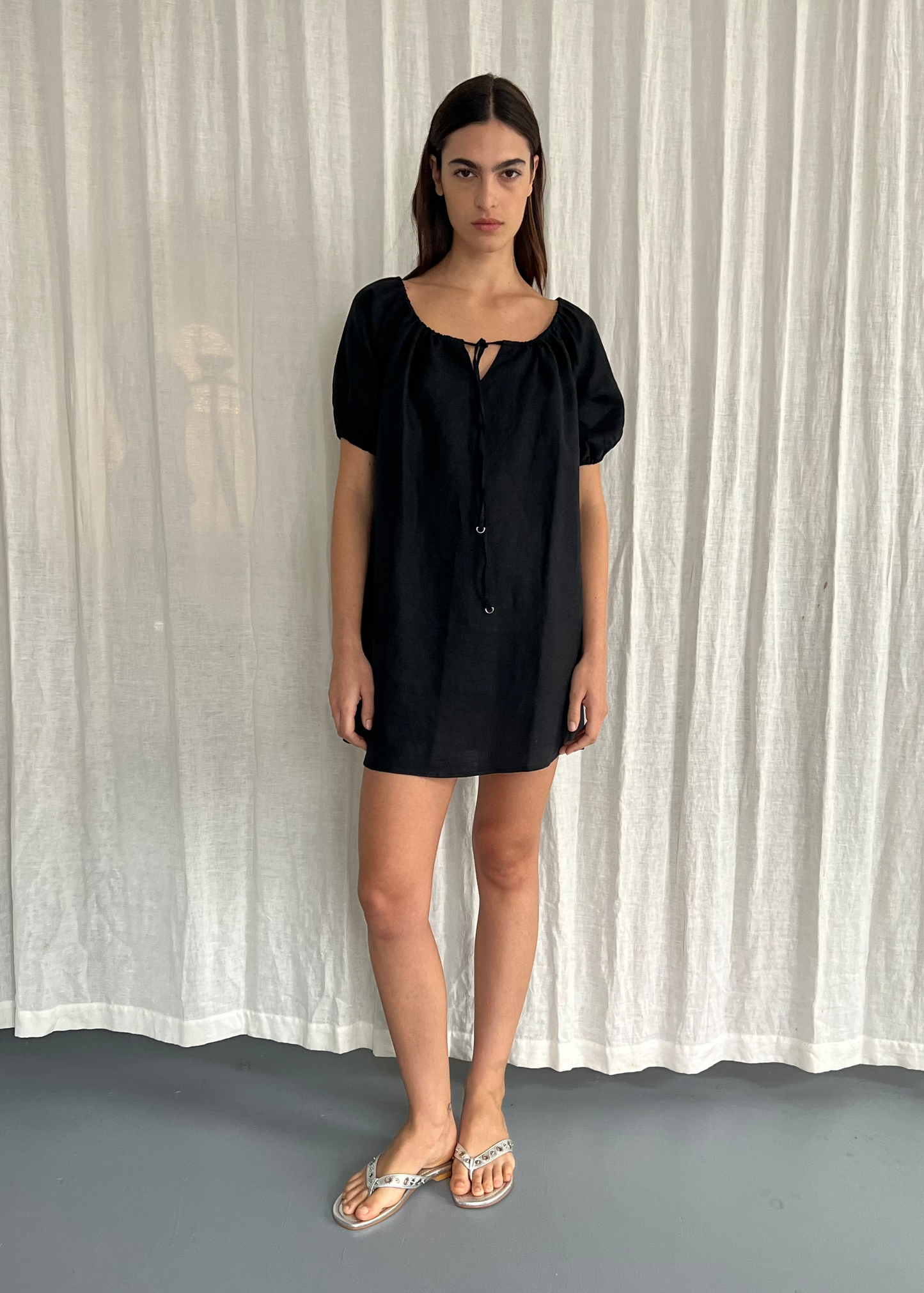 A woman with straight brown hair and a neutral expression stands before sheer white curtains, wearing the Smock Dress - Black by Deiji Studios, which features short puff sleeves and a tie neckline, paired with silver sandals.