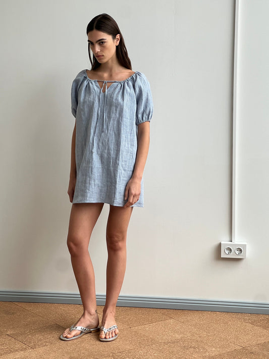 A woman stands indoors near a plain wall wearing the Smock Dress - Blue Stripe, featuring subtle stripes and a front tie detail, paired with silver sandals. Two electrical outlets are visible in the background.