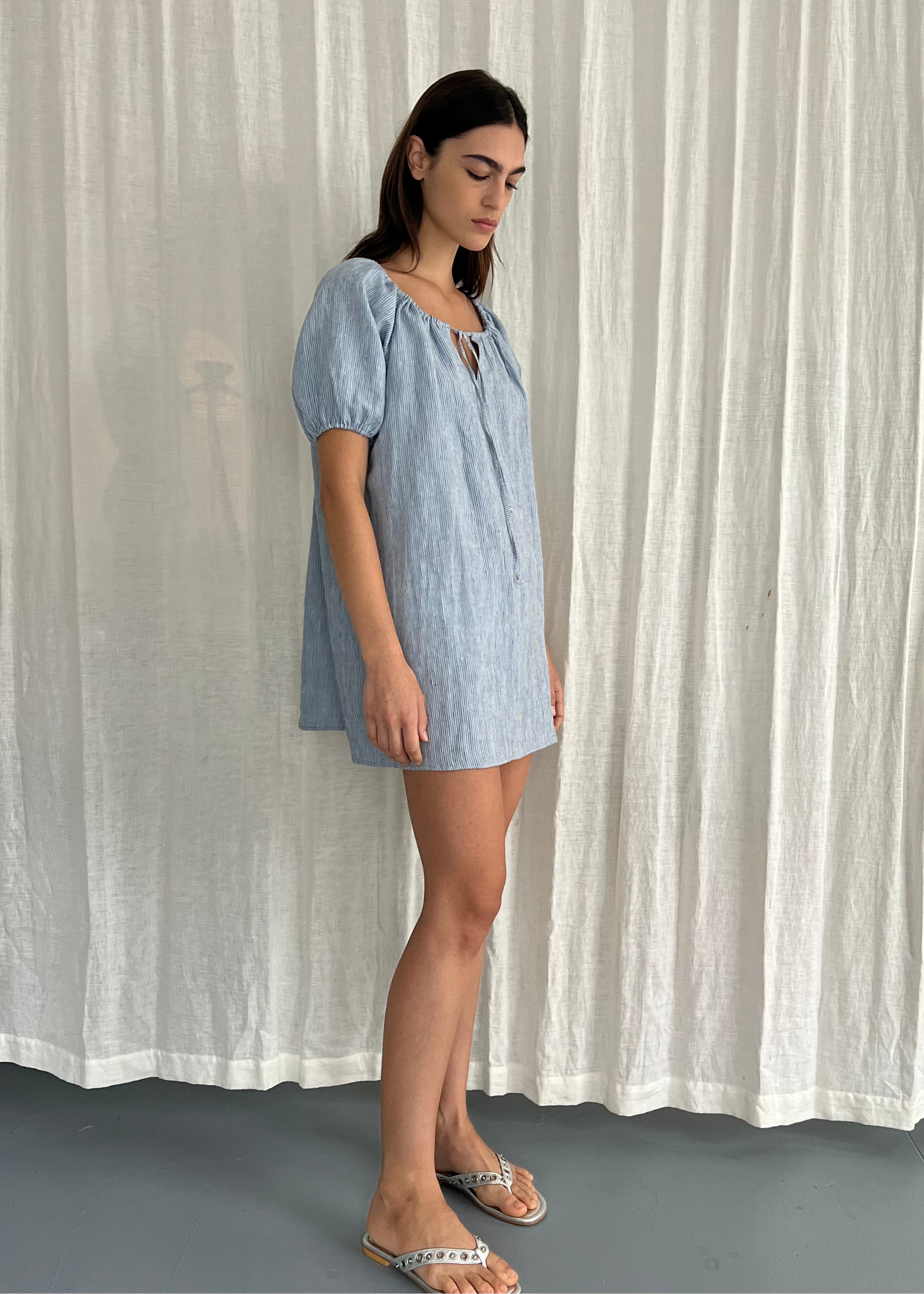 A woman stands in profile wearing the Smock Dress - Blue Stripe, a light blue, short-sleeved, loose-fitting dress paired with flat sandals, set against a white textured curtain background.