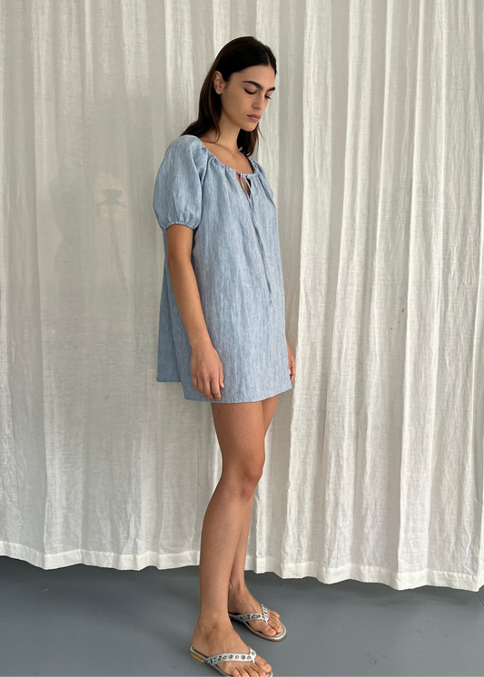 A woman stands in profile wearing the Smock Dress - Blue Stripe, a light blue, short-sleeved, loose-fitting dress paired with flat sandals, set against a white textured curtain background.