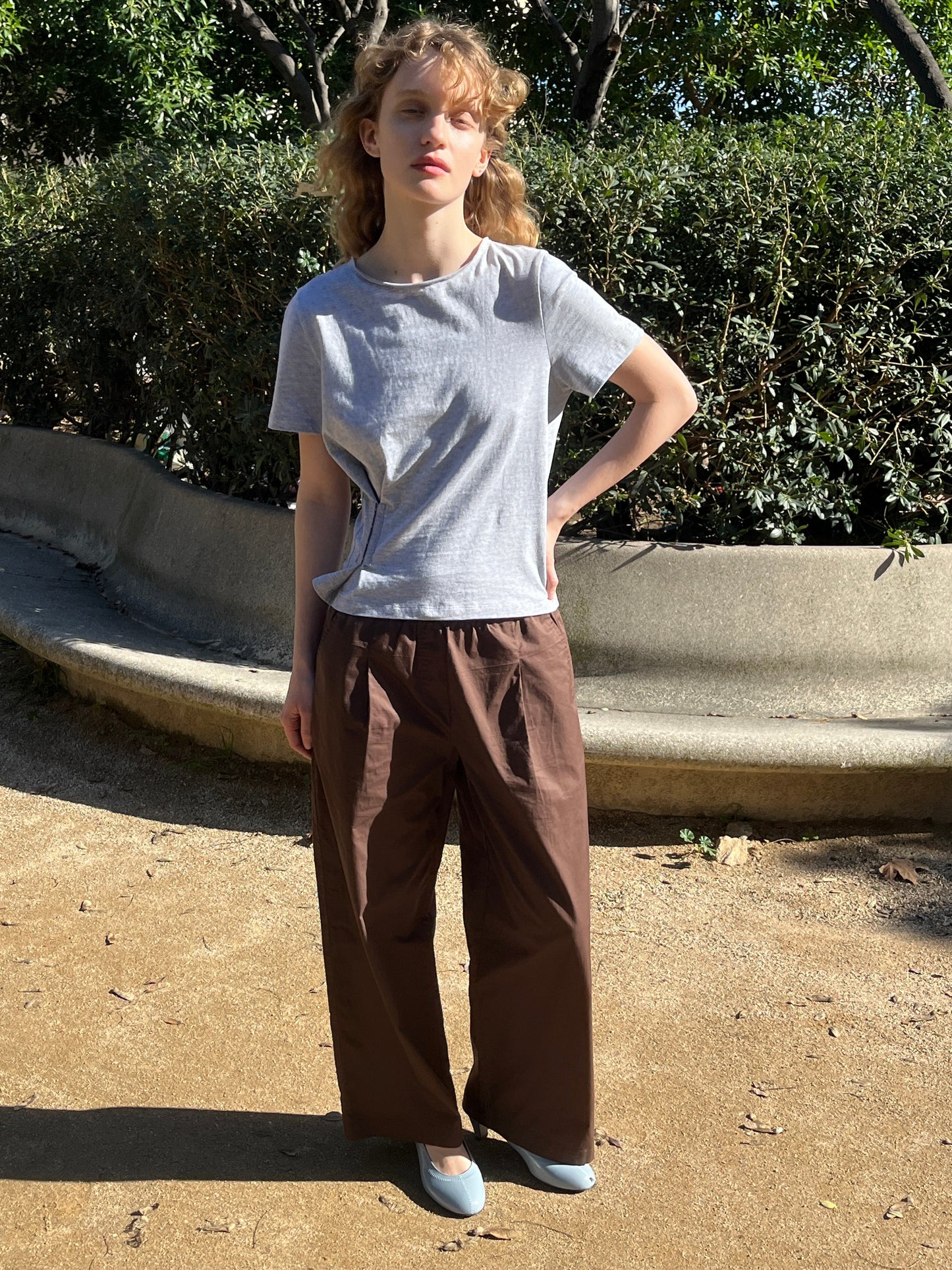 A young woman with curly blonde hair stands outdoors on a sandy path, wearing the Contrast Waist Pant - Brown by Deiji Studios, a light gray T-shirt, and light blue shoes. Green bushes and a curved concrete bench are in the background.