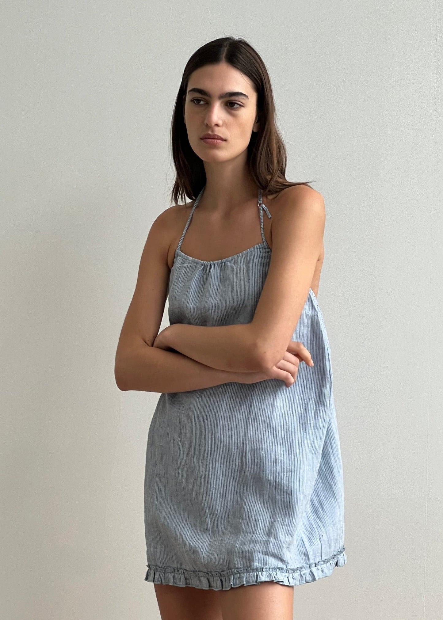 A woman with long brown hair stands against a light wall, wearing Deiji Studios’ Flounce Halter Dress - Blue Stripe. The sleeveless dress features thin straps and a ruffled hem; she stands with crossed arms and a neutral expression.