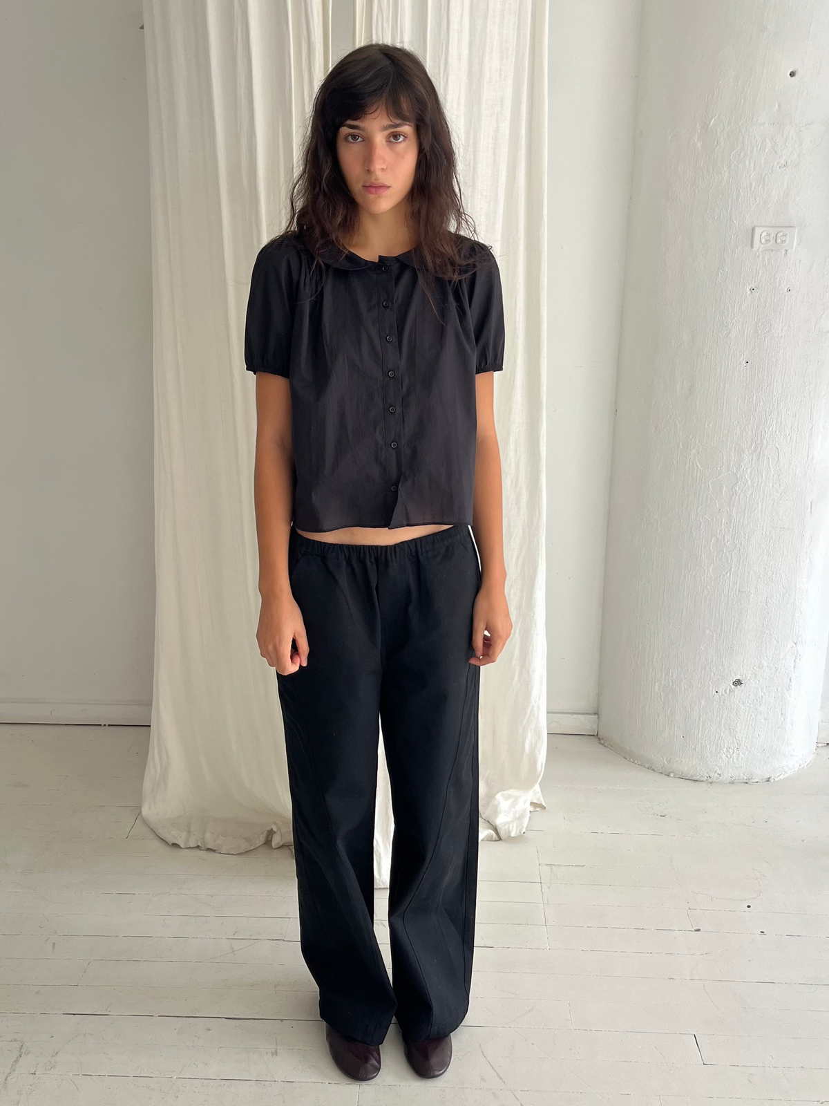 A woman with dark wavy hair stands against a white curtain, wearing the Curve Pant - Black by Deiji Studios, a softly structured pant with a relaxed fit and subtle curved leg, styled with the Petal Top in Black.