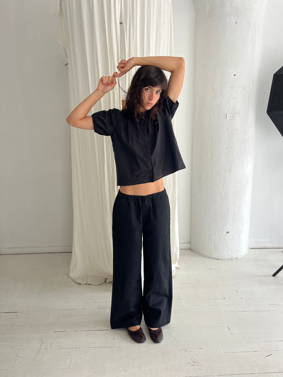 A woman stands against a white curtain with arms raised, wearing the Curve Pant - Black by Deiji Studios, a softly structured pant with a relaxed fit and subtle curved leg, styled with the Petal Top in Black.