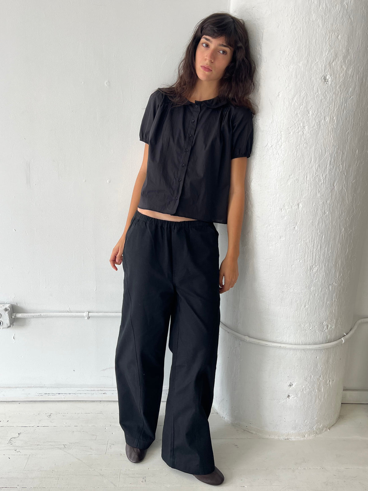 A woman leans against a white column, wearing the Curve Pant - Black by Deiji Studios, a softly structured pant with a relaxed fit and subtle curved leg, styled with the Petal Top in Black.