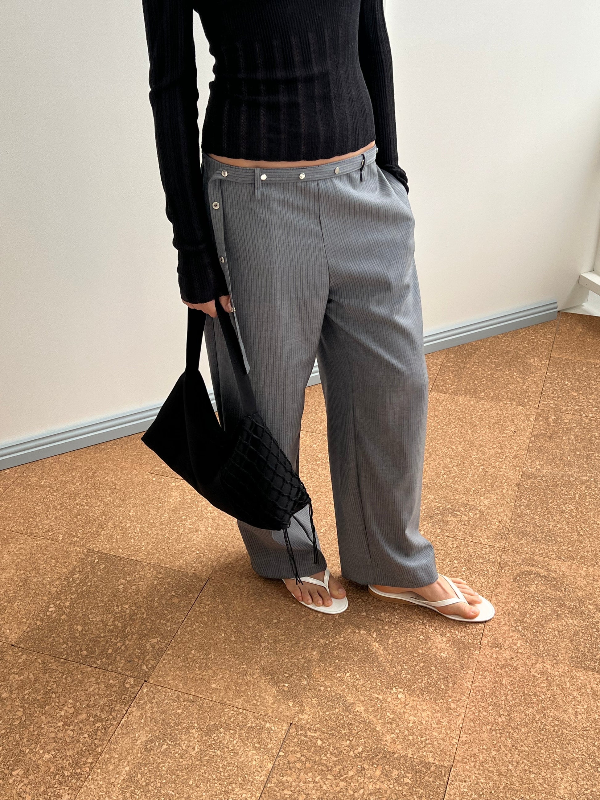 A person stands indoors wearing gray pinstripe pants, a black ribbed long-sleeve top, white flip-flops, and carries the Net Contrast Bag - Black. Only their torso and legs are visible.