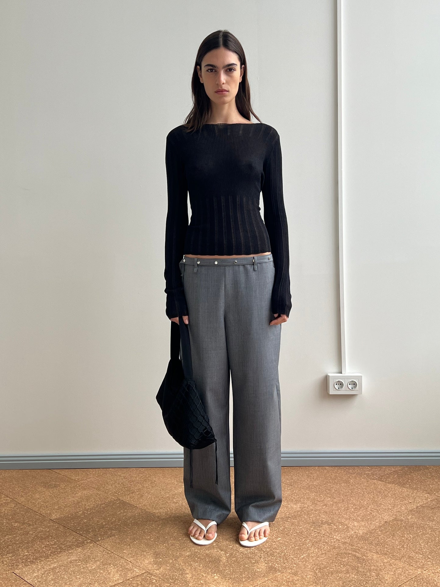 A woman stands indoors against a plain wall, wearing a fitted black long-sleeve top, gray wide-leg trousers with a brown waistband, white sandals, and holding the Net Contrast Bag - Black from Deiji Studios.