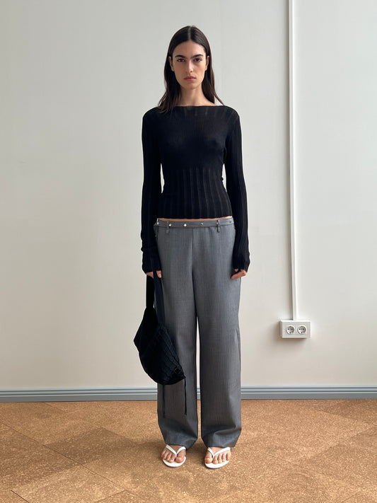 A woman stands indoors against a plain wall, wearing a fitted black long-sleeve top, gray wide-leg trousers with a brown waistband, white sandals, and holding the Net Contrast Bag - Black from Deiji Studios.