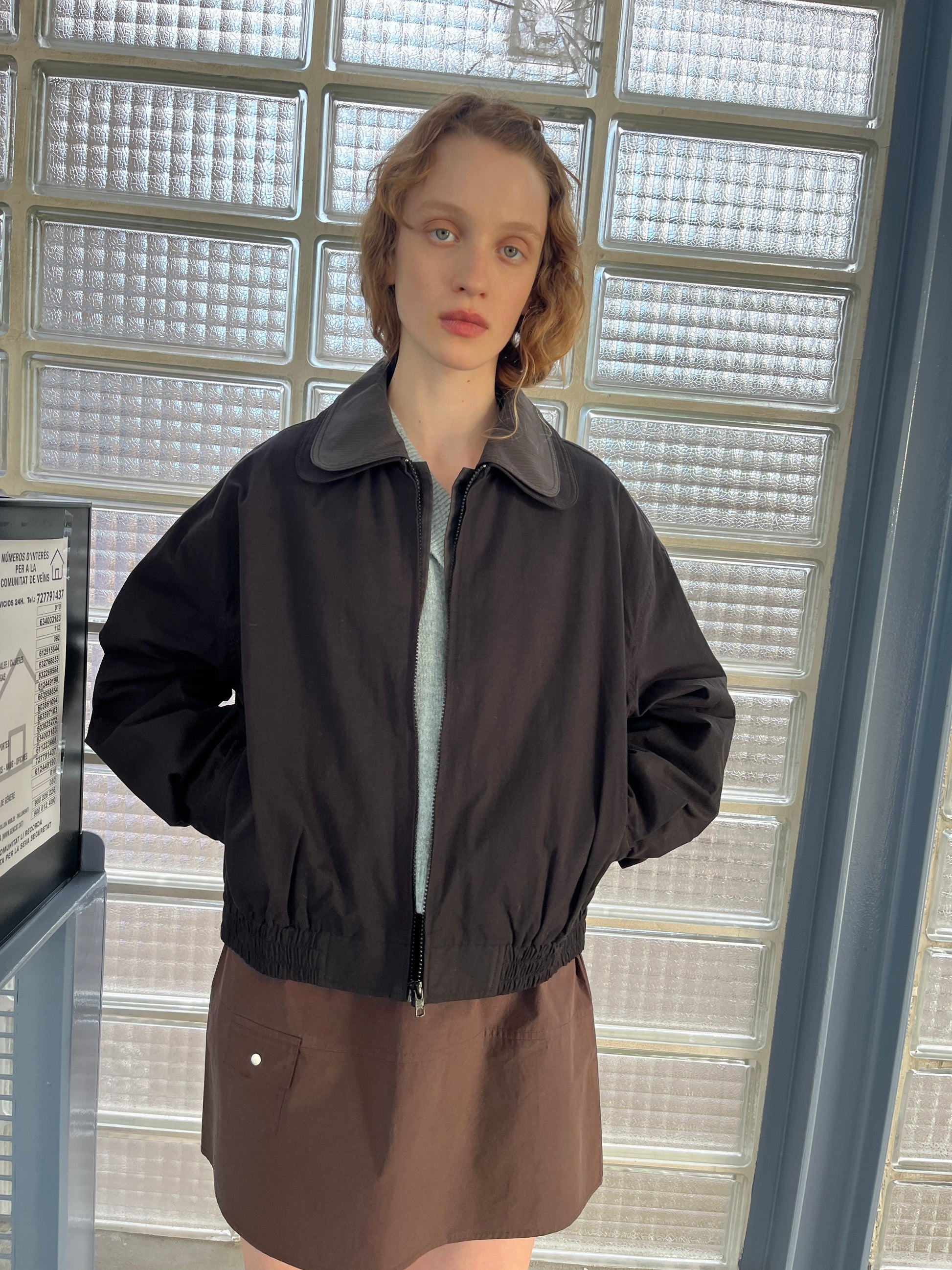 A person with short, wavy hair stands indoors by glass block windows, wearing the Collared Zip Jacket - Black Contrast over a light top and brown skirt, looking directly at the camera with a neutral expression.