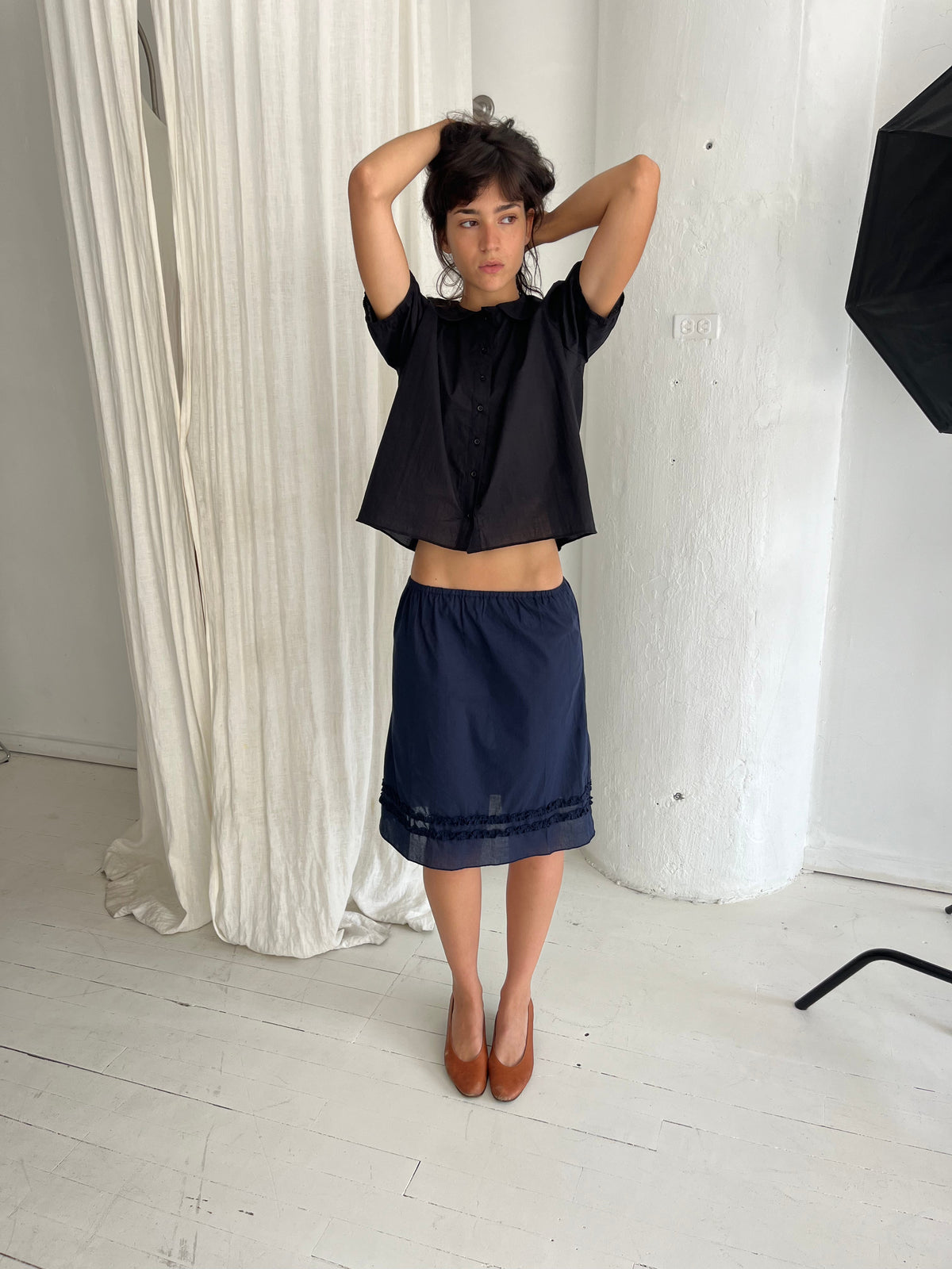 A woman stands against a white curtain with arms raised, wearing the Daytime Skirt - Navy by Deiji Studios, a cotton voile midi skirt with double frill detailing at the hem, styled with the Petal Top in Black.