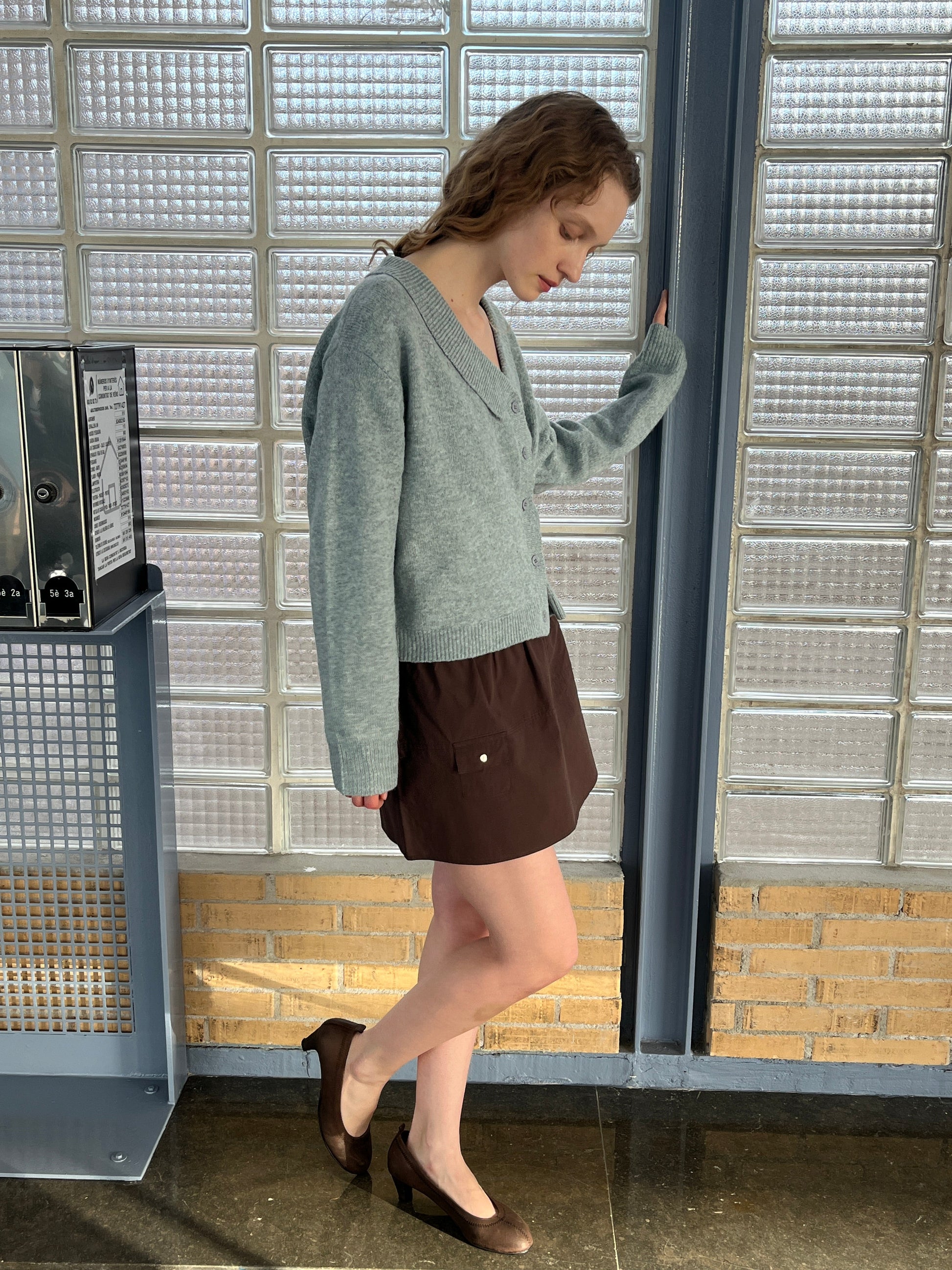 A woman stands indoors near glass bricks, looking down with one hand on a metal beam. She wears a light blue cardigan, the Pocket Skirt in Sepia by Deiji Studios, and brown heels. Sunlight casts shadows on the floor and wall behind her.
