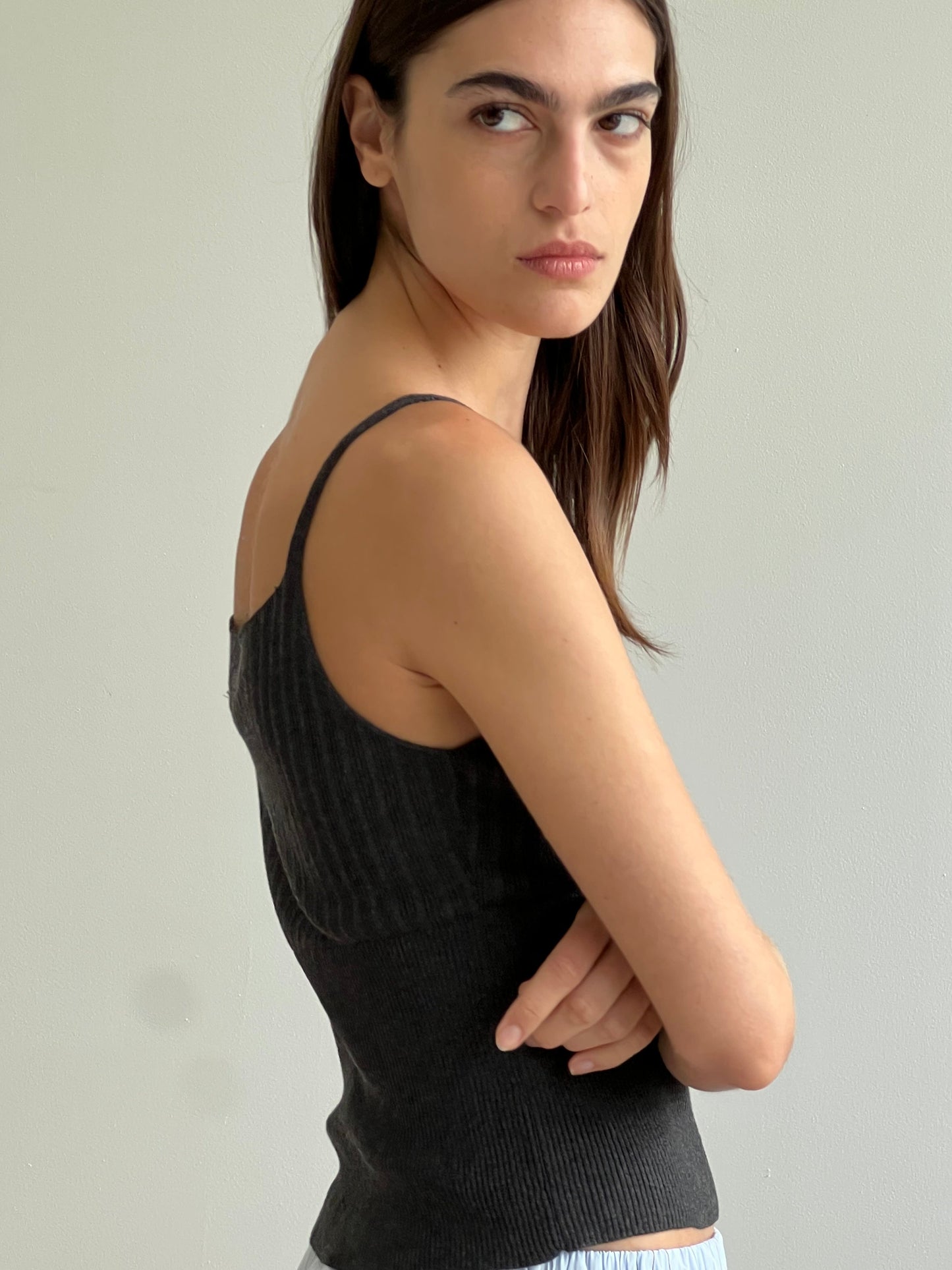 A woman with long brown hair wears the Ribbed Tank - Dark Charcoal by Deiji Studios, standing sideways against a plain light wall and looking over her shoulder with a serious expression.