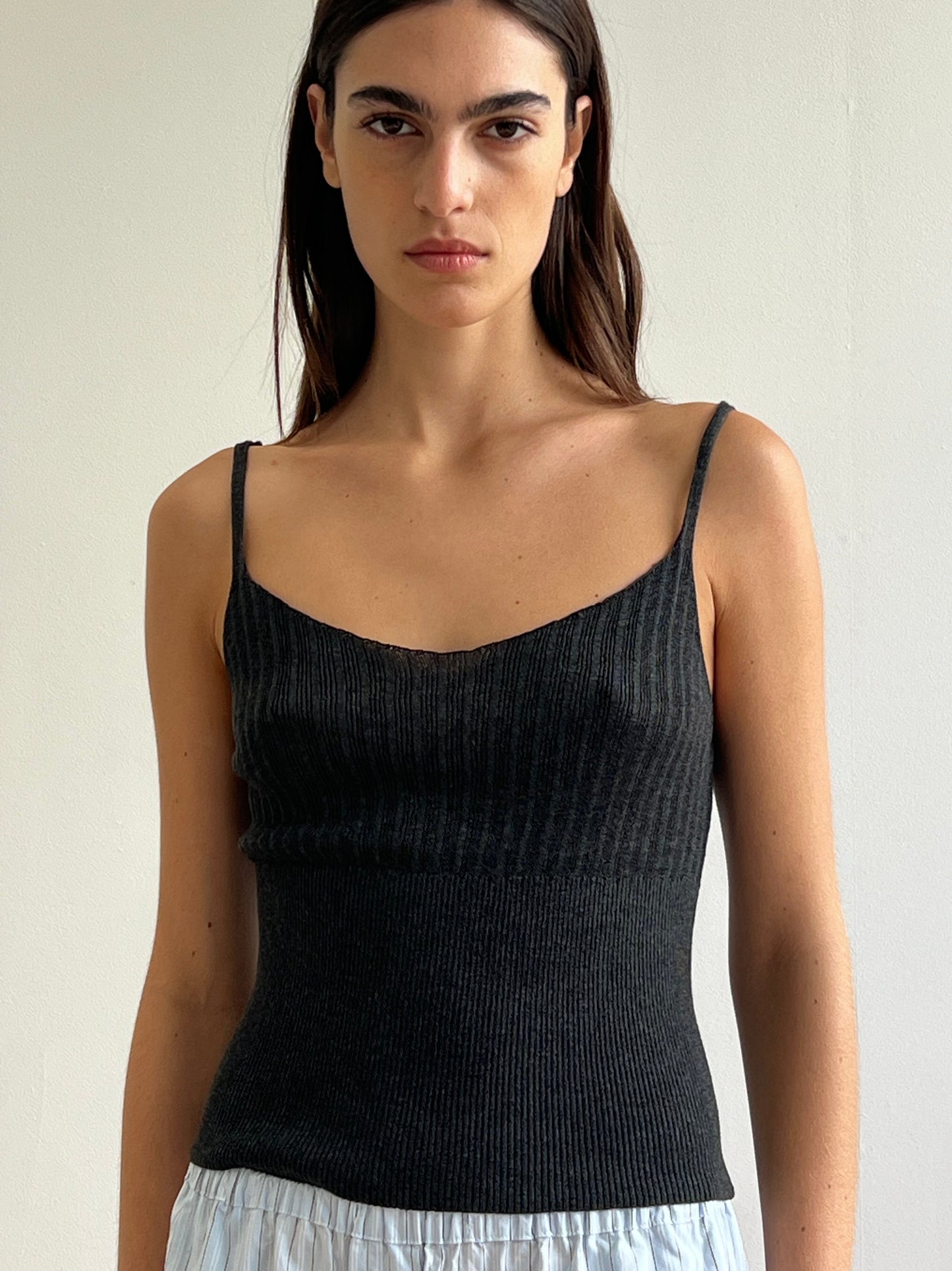 A woman with long brown hair wears Deiji Studios' Ribbed Tank in Dark Charcoal and light blue pleated pants, standing against a plain light background and looking neutrally at the camera.