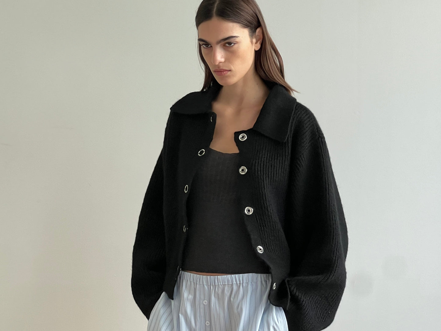 A woman with straight brown hair wears the Stud Cardi Knit - Black, an oversized sustainable cardigan, over a black top and light blue pleated skirt, standing against a plain light-colored wall.