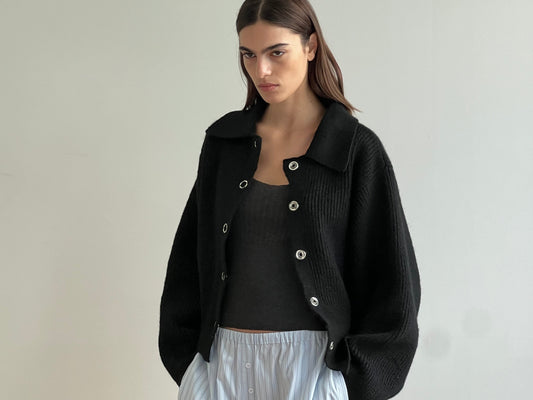A woman with straight brown hair wears the Stud Cardi Knit - Black, an oversized sustainable cardigan, over a black top and light blue pleated skirt, standing against a plain light-colored wall.