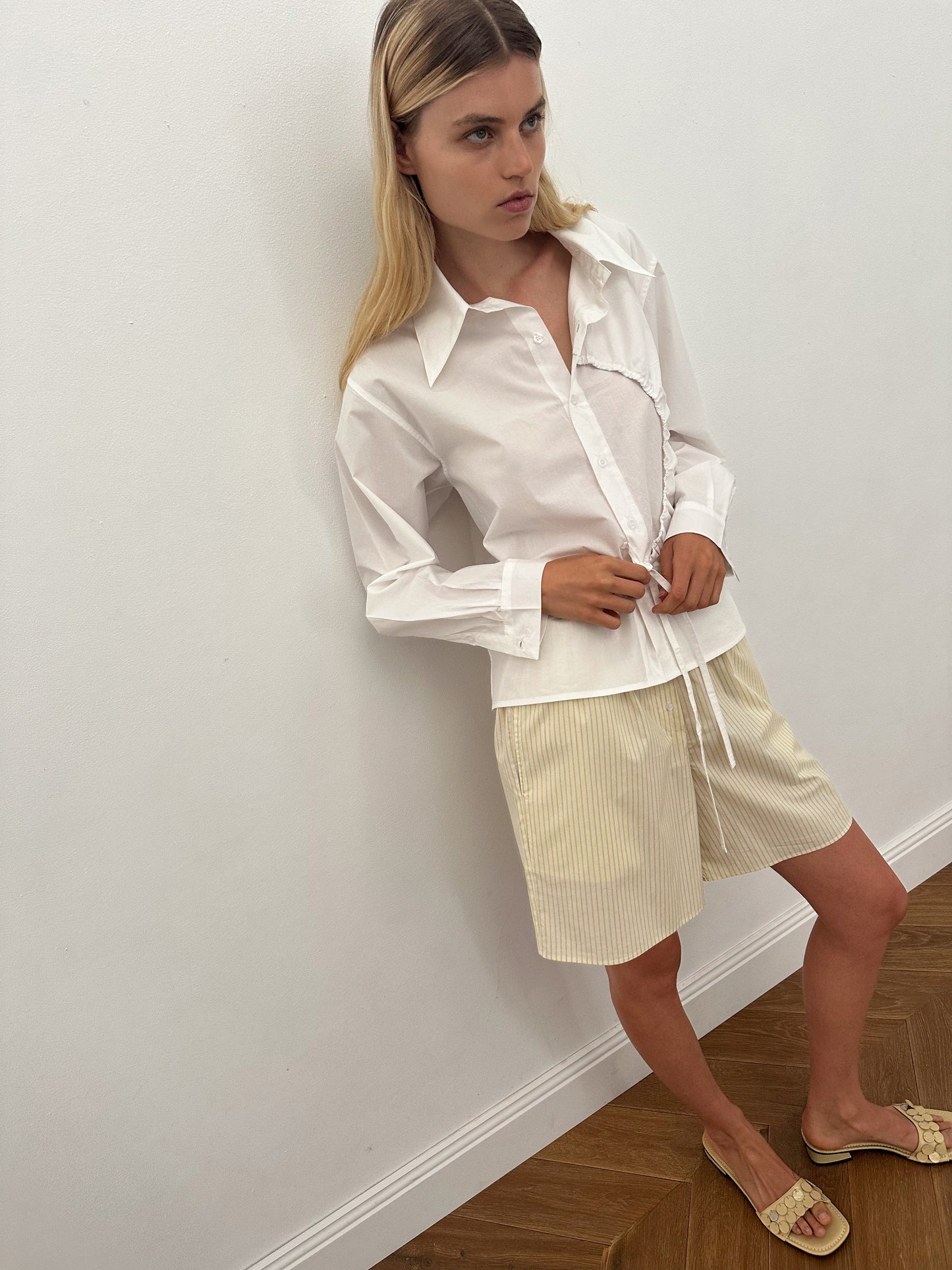 A woman with long blonde hair wears the Arch Tie Shirt - White by Deiji Studios, styled with beige pinstriped shorts. She stands indoors on a wooden floor, in front of a plain white wall, looking off to the side in a relaxed pose.