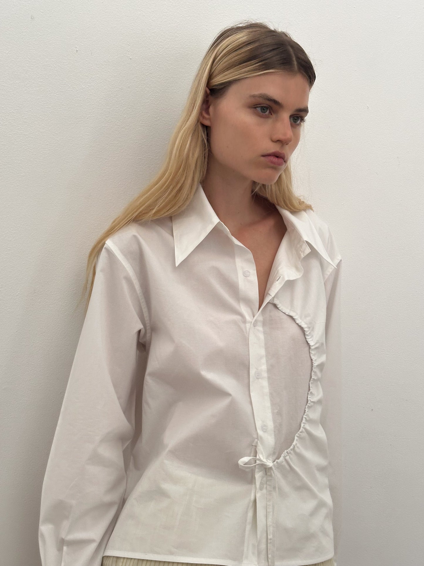 A woman with long blonde hair wears the Arch Tie Shirt - White by Deiji Studios, a boxy fit button-up featuring an asymmetrical cut-out, standing against a plain white wall and gazing to the side with a neutral expression.