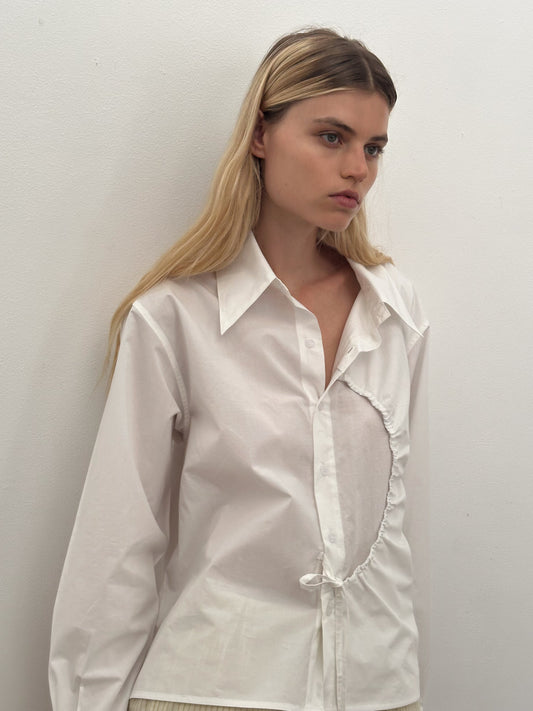 A woman with long blonde hair wears the Arch Tie Shirt - White by Deiji Studios, a boxy fit button-up featuring an asymmetrical cut-out, standing against a plain white wall and gazing to the side with a neutral expression.