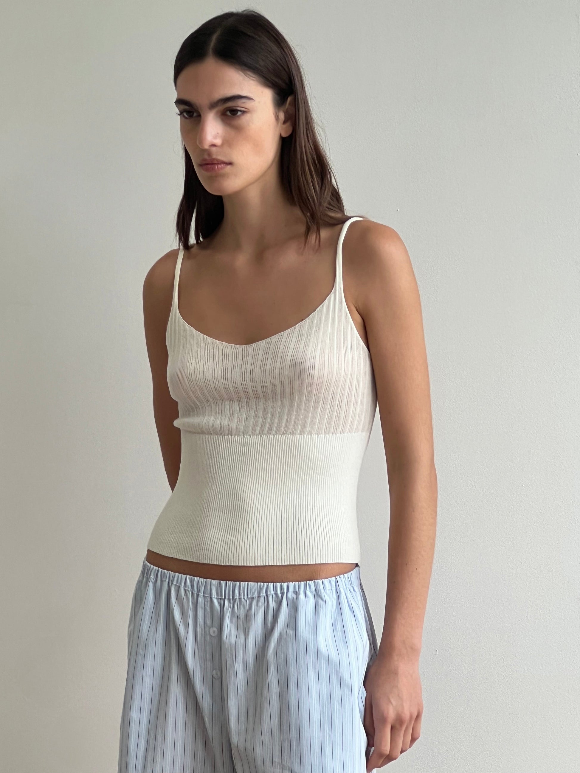 A woman with long dark hair stands against a light wall, wearing the Deiji Studios Ribbed Tank - Off White and light blue striped lyocell pants. Her arms are relaxed at her sides and she has a neutral expression.