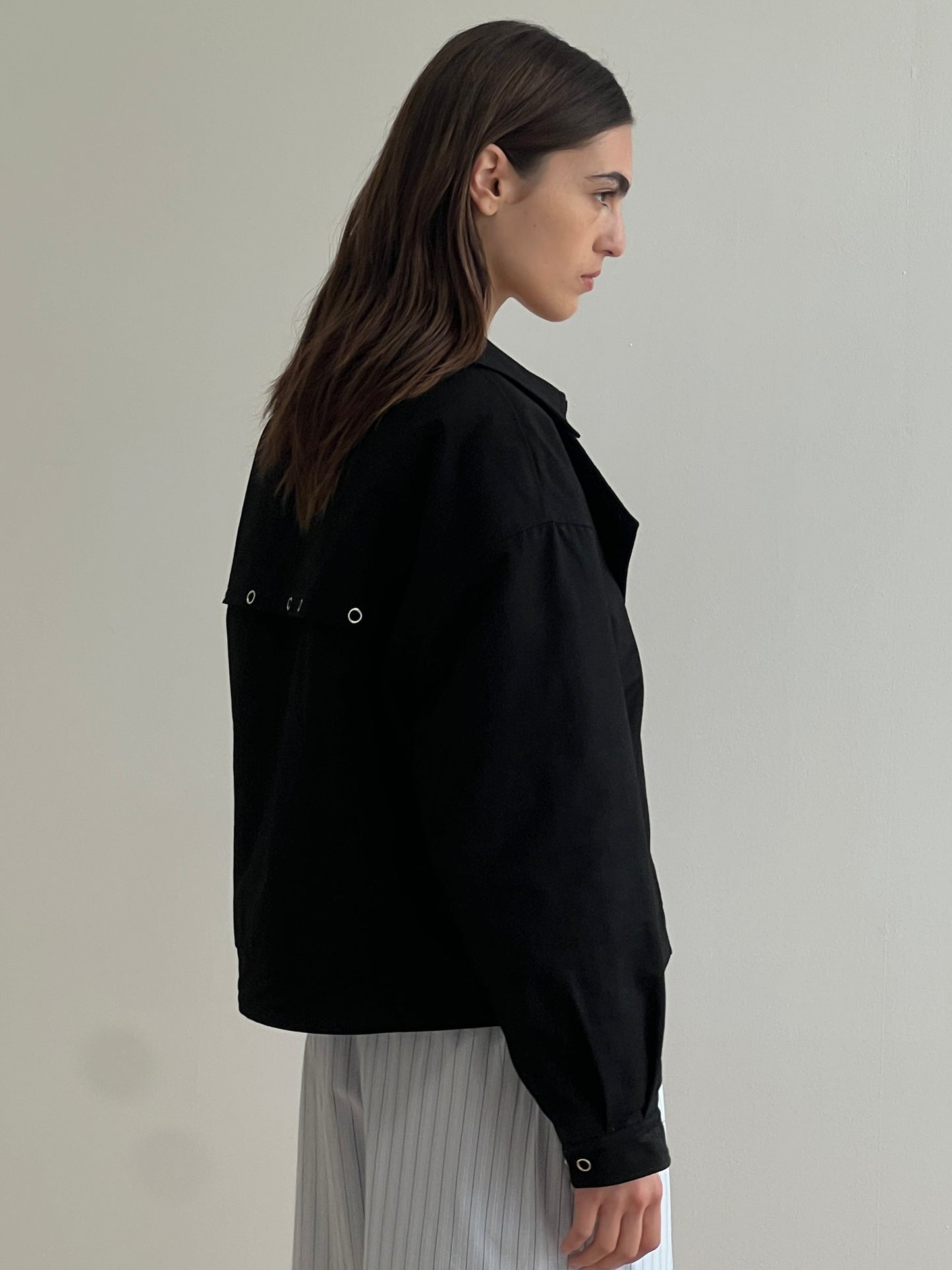 A woman with long brown hair stands sideways, wearing the Lapel Jacket - Black by Deiji Studios, which features silver grommets on the back, paired with light-colored vertically striped pants against a plain light background.