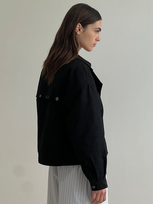 A woman with long brown hair stands sideways, wearing the Lapel Jacket - Black by Deiji Studios, which features silver grommets on the back, paired with light-colored vertically striped pants against a plain light background.