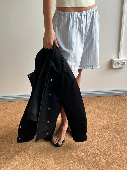 A person in a white tank top, blue-and-white striped shorts, and sandals holds the Deiji Studios Lapel Jacket - Black by their side while standing on a brown floor near a white wall with power outlets.