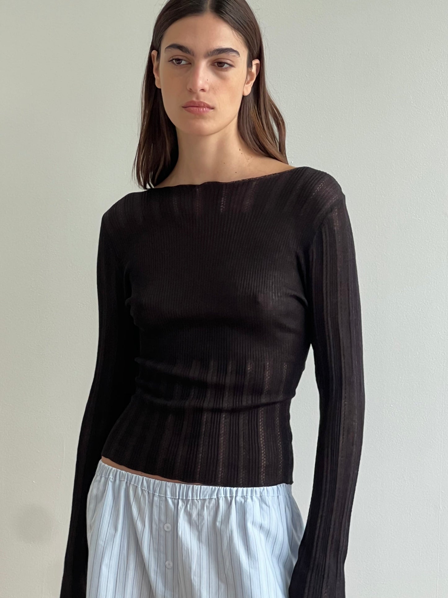 A woman with straight brown hair wears the Open Work Longsleeve - Black from Deiji Studios and loose, light blue striped pants, standing against a plain light background and looking slightly to the side with a neutral expression.
