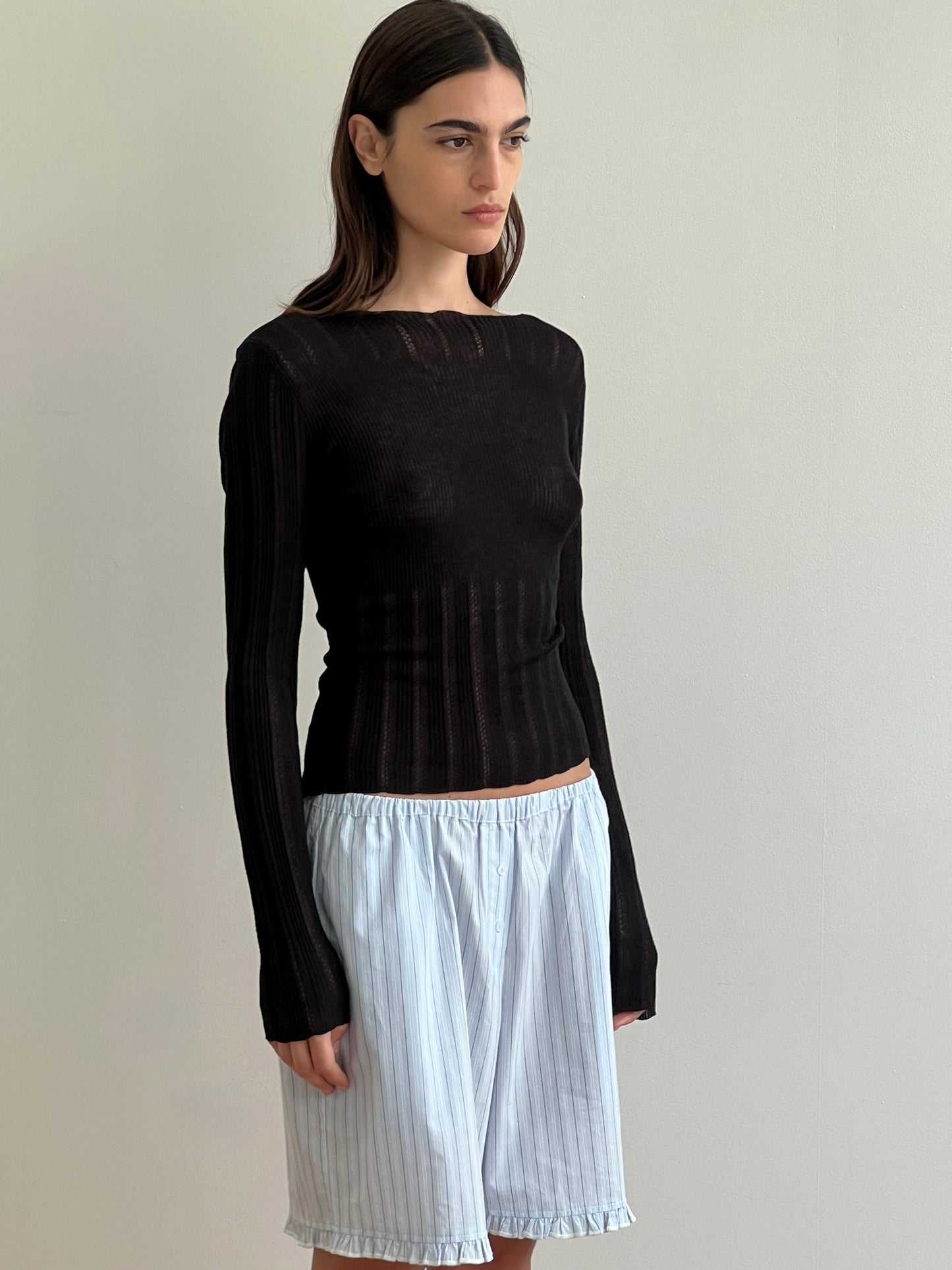 A woman with long dark hair wears the Open Work Longsleeve in black by Deiji Studios and loose, knee-length pinstriped white shorts, standing against a plain light-colored wall.