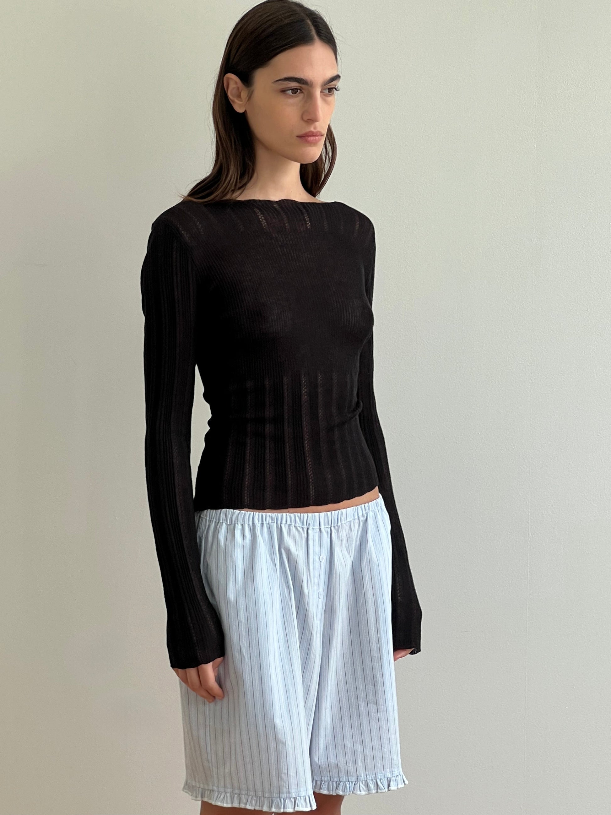 A woman with straight brown hair stands against a light wall, wearing Deiji Studios' Open Work Longsleeve - Black paired with loose white shorts featuring thin vertical stripes.