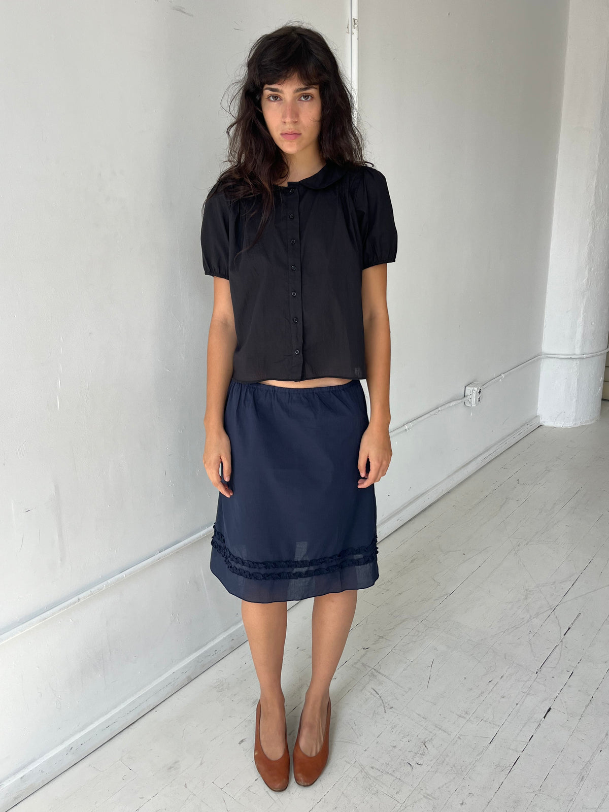 A woman with dark wavy hair stands against a white wall, wearing the Daytime Skirt - Navy by Deiji Studios, a cotton voile midi skirt with a straight silhouette and double frill detailing at the hem, styled with the Petal Top in Black.