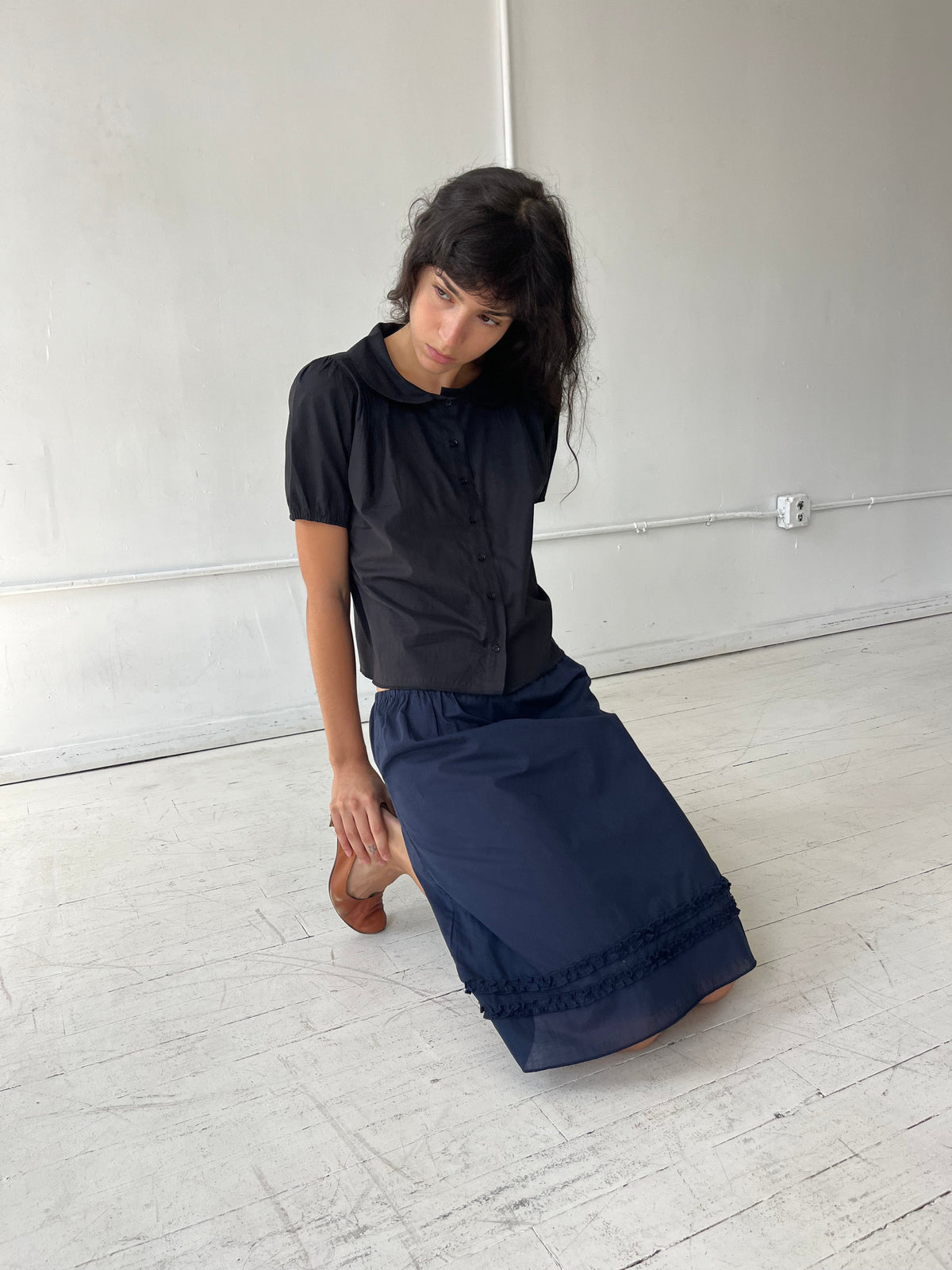 A woman kneels on a white studio floor, wearing the Daytime Skirt - Navy by Deiji Studios, a cotton voile midi skirt with double frill detailing at the hem, styled with the Petal Top in Black.