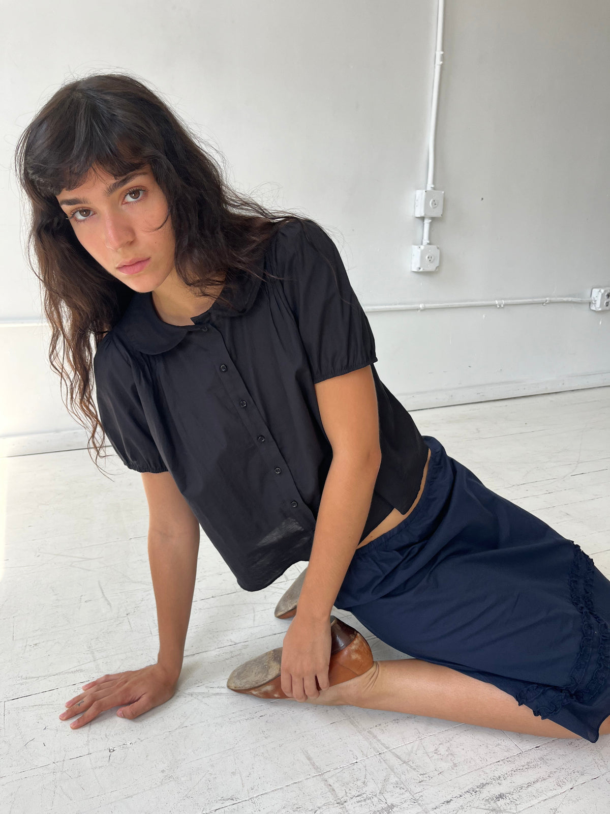 A woman sits on a white studio floor, wearing the Petal Top - Black by Deiji Studios, a cotton voile top with a peter pan collar and puffed short sleeves, styled with the Daytime Skirt in Navy.