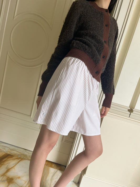A person stands indoors wearing the Button Short - Musk Stripe, a relaxed fit brown button-up cardigan, and a white skirt with subtle vertical stripes. The cream paneled walls and marble floor complete the background; the face is not visible.