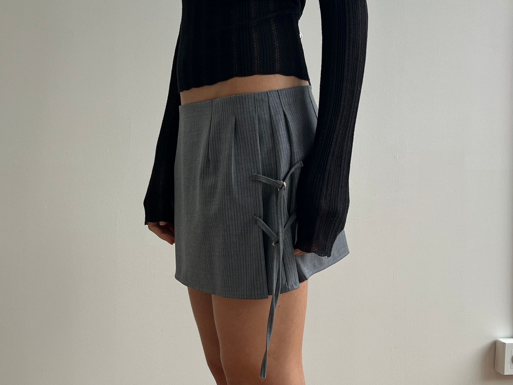 Person wearing a black long-sleeve top and a Tab Tie Skirt - Grey Blue Stripe by Deiji Studios, featuring side lace-up detail, standing against a plain white wall with a visible electrical outlet and pipe.