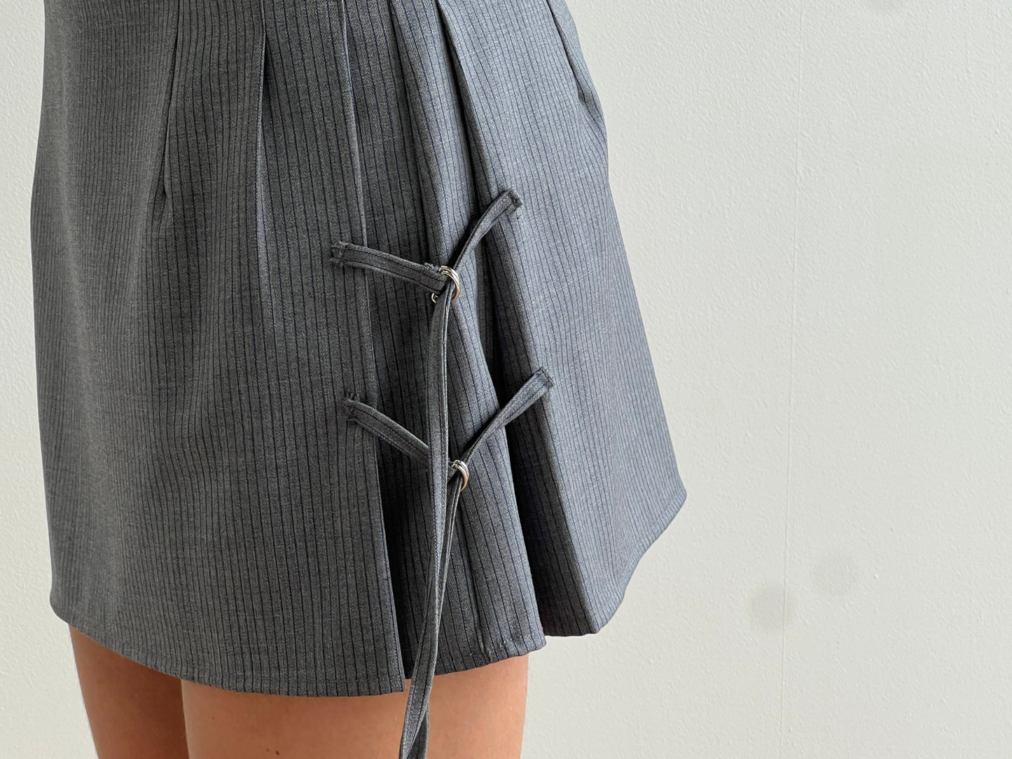A close-up of a person wearing the Tab Tie Skirt - Grey Blue Stripe by Deiji Studios, featuring decorative crisscross laces on the side, crafted from soft RWS wool, set against a plain white background.