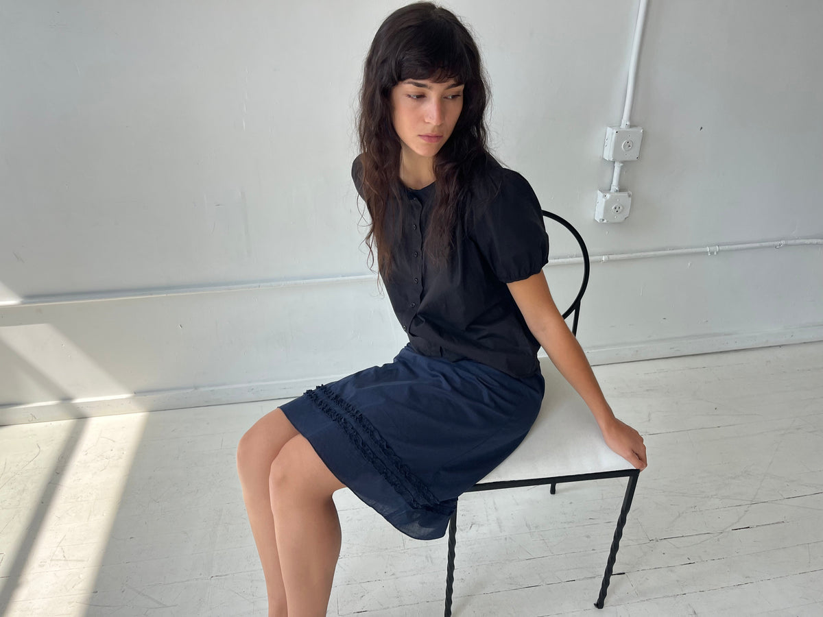 A woman sits on a white bench in a studio, wearing the Daytime Skirt - Navy by Deiji Studios, a cotton voile midi skirt with double frill detailing at the hem, styled with the Petal Top in Black.