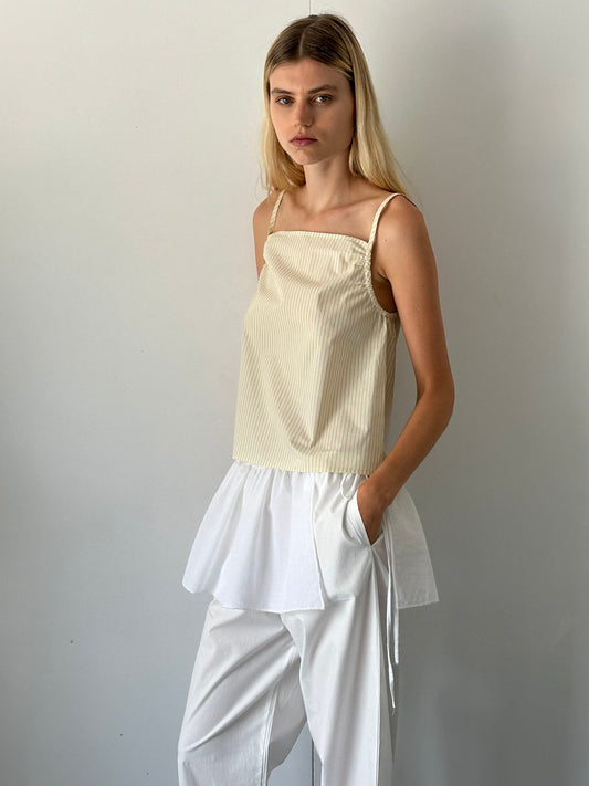 A woman with long blonde hair stands against a white wall, wearing the Gathered Strap Top - Butter Stripe by 0 and loose white pants. She has a neutral expression and her hands in her pockets.