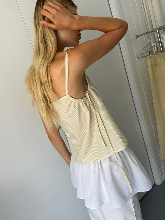 A woman with long blonde hair stands sideways near a clothing rack, touching her head. She wears the Gathered Strap Top - Butter Stripe by 0 in organic cotton poplin over a white blouse and white pants indoors.